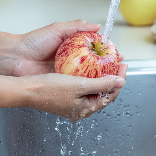 Persona lavando una manzana bajo un chorro de agua para garantizar la higiene alimentaria y eliminar residuos superficiales.